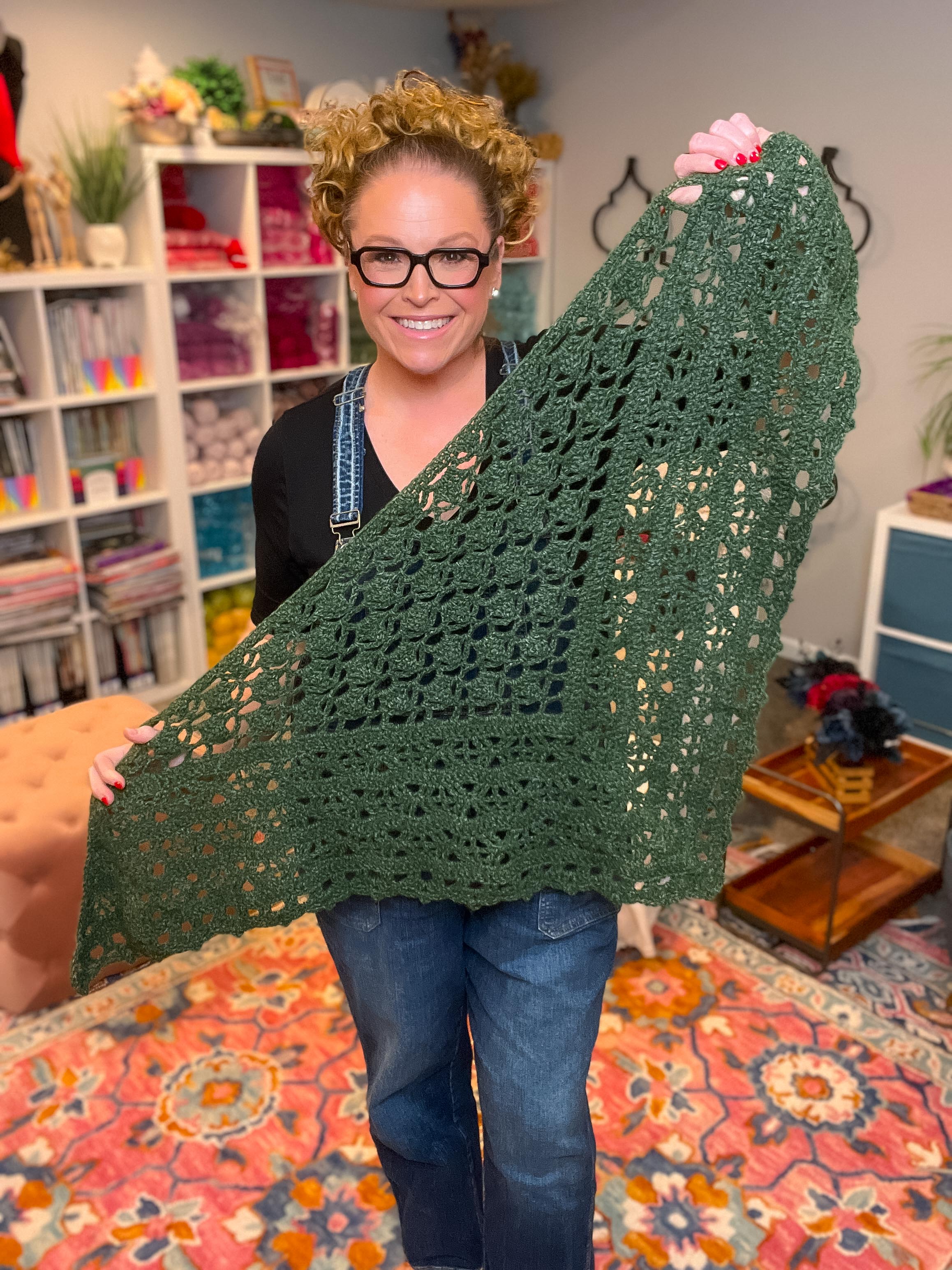 Person holding a green crocheted shawl in a room with shelves and decor.