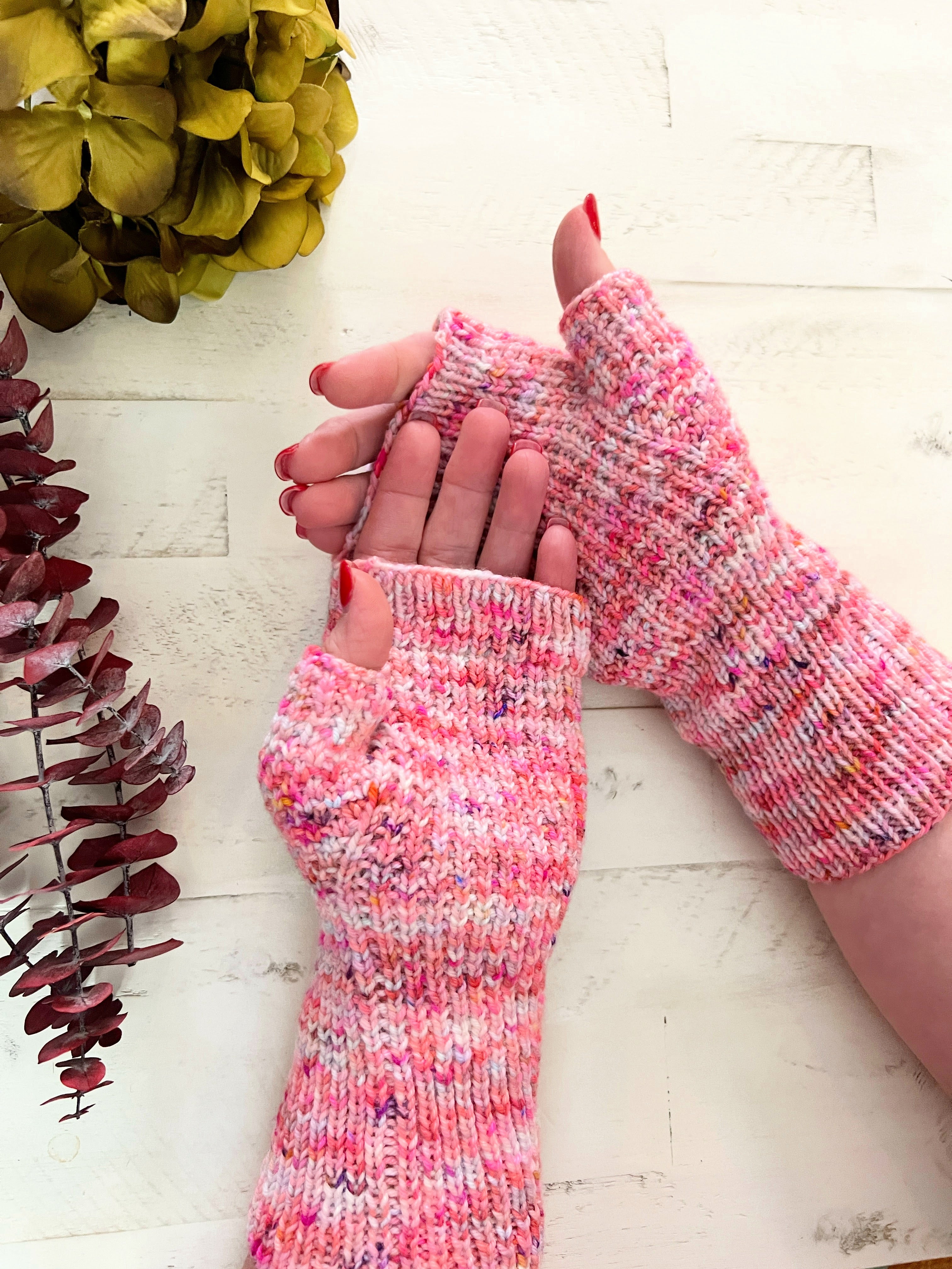 Pink speckled knit fingerless mittens showing textured hands, shaped thumbs, and long ribbed cuffs against white wood.