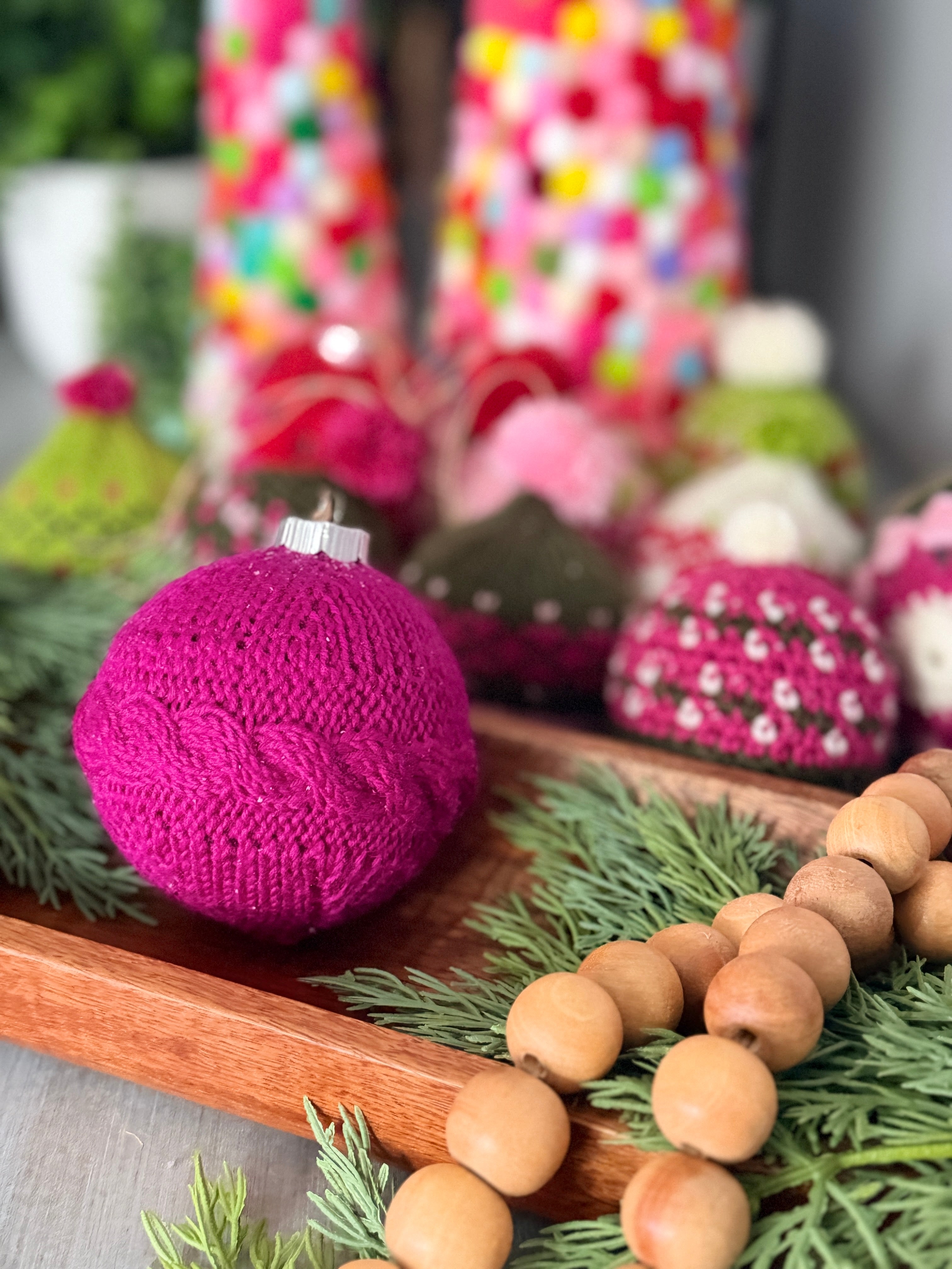 A round ornament created using the “My Tree Needed This Knit Ornament Pattern PDF - Knit round ornament Flat” in magenta sits on a wooden tray with greenery and wooden beads, surrounded by colorful, knit holiday decorations in the background.