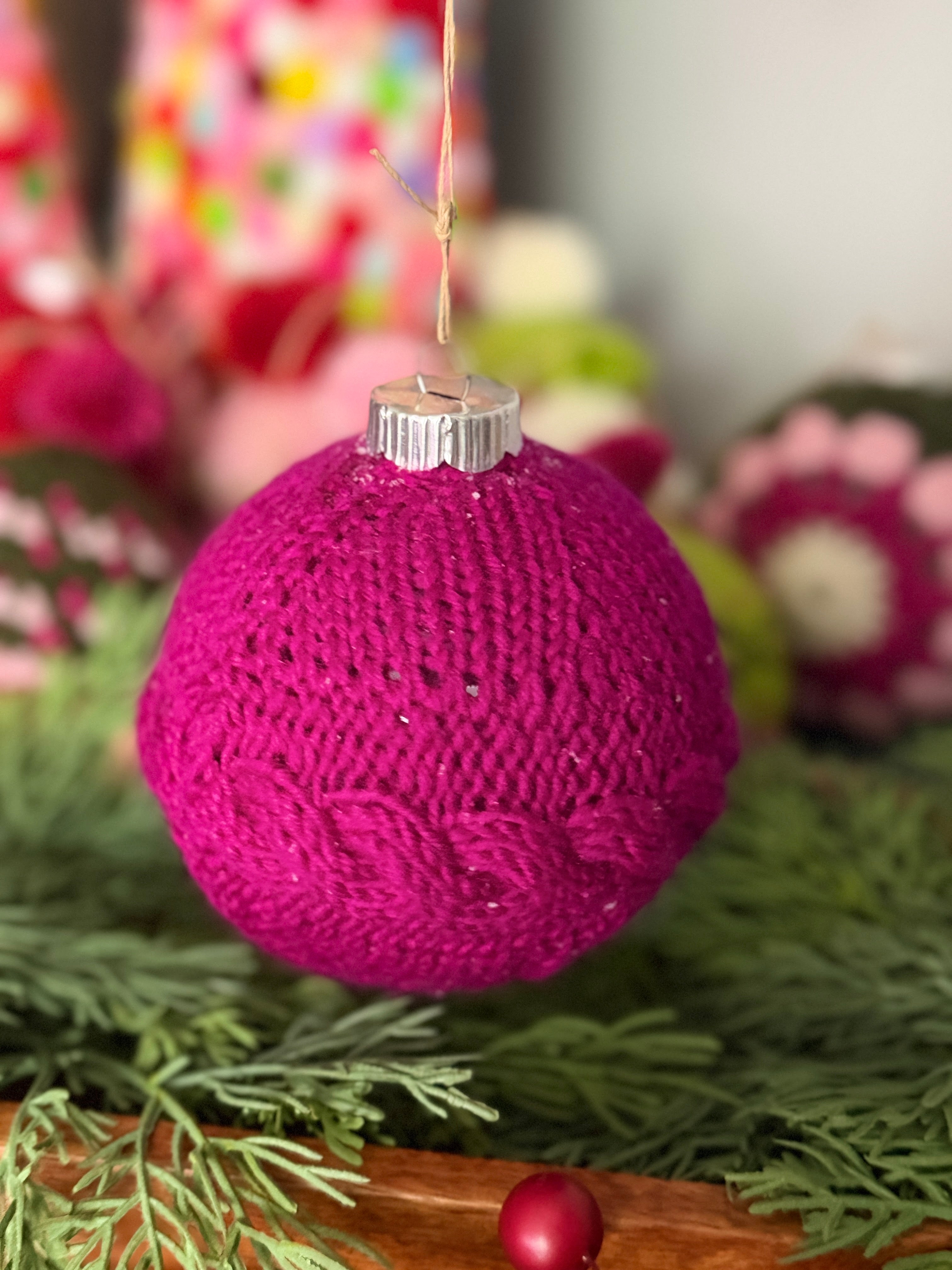 A festive, purple knitted ornament made using the “My Tree Needed This Knit Ornament Pattern PDF - Knit round ornament Flat” hangs from a string, with vibrant greenery and other colorful holiday decorations softly blurred in the background.