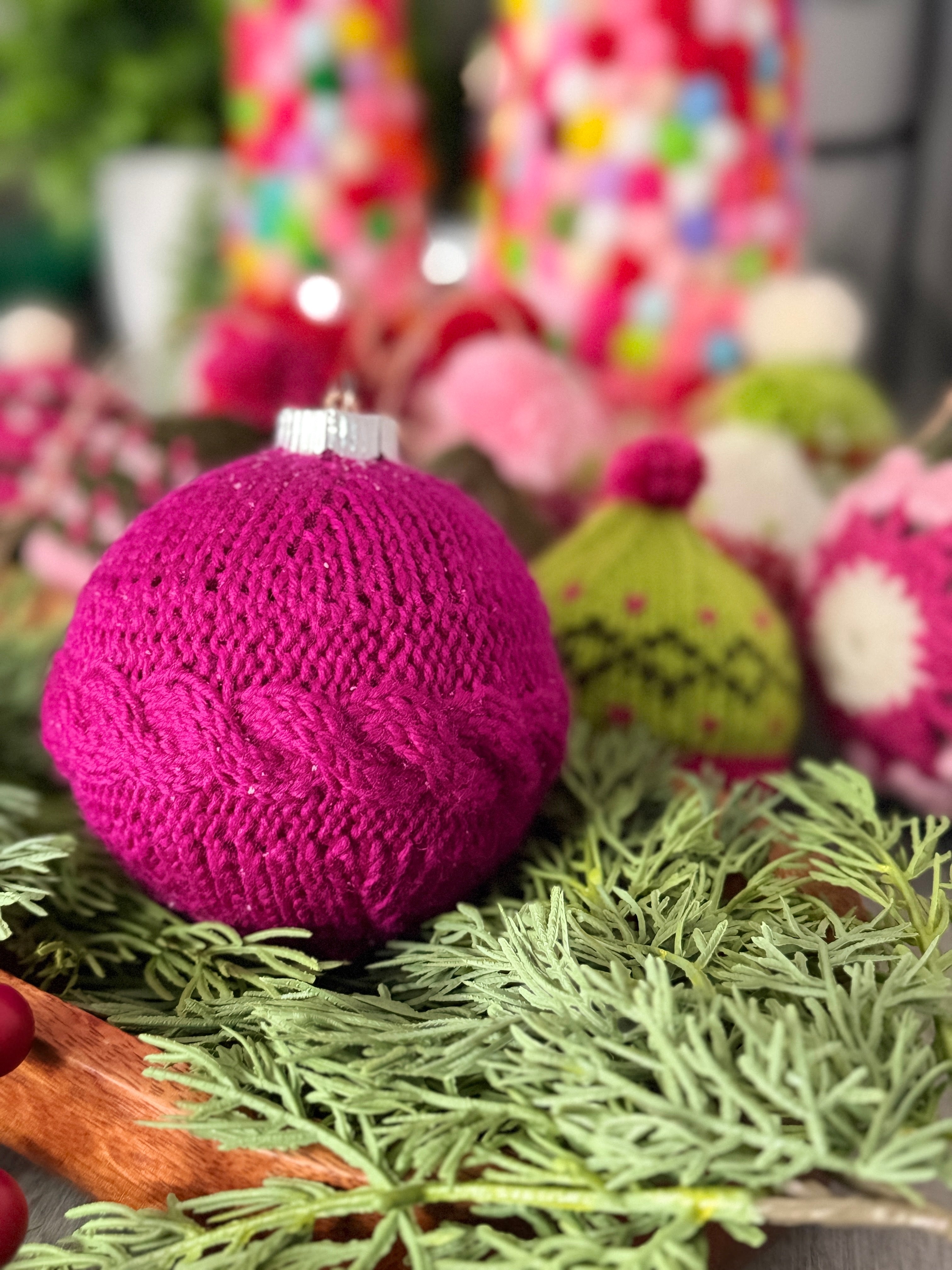 A vibrant magenta Christmas ornament, created with the *My Tree Needed This Knit Ornament Pattern PDF - Knit round ornament Flat*, features a classic cable pattern and rests among festive greenery, surrounded by other colorful knitted holiday decorations—perfect for displaying your favorite knit design or adding a signature Marly Bird touch to your seasonal décor.